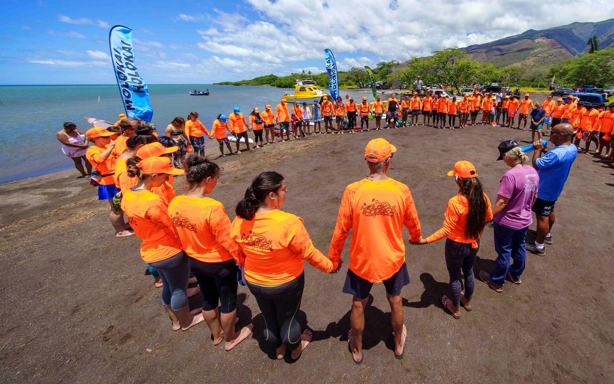 Participants in orange shirts hold hands in a large circle by the ocean
